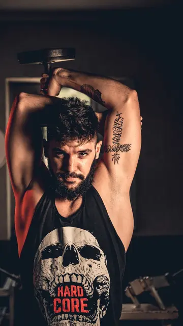 Placement still — Bearded man with tattoos exercising by lifting a dumbbell overhead in a gym setting.