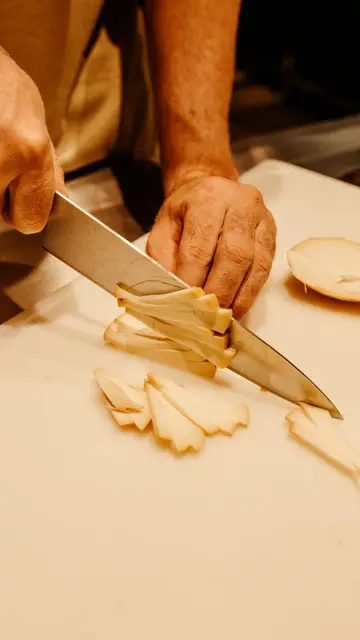 Podcasts placement — A close-up view showing hands slicing a vegetable on a white cutting board using a sharp knife.